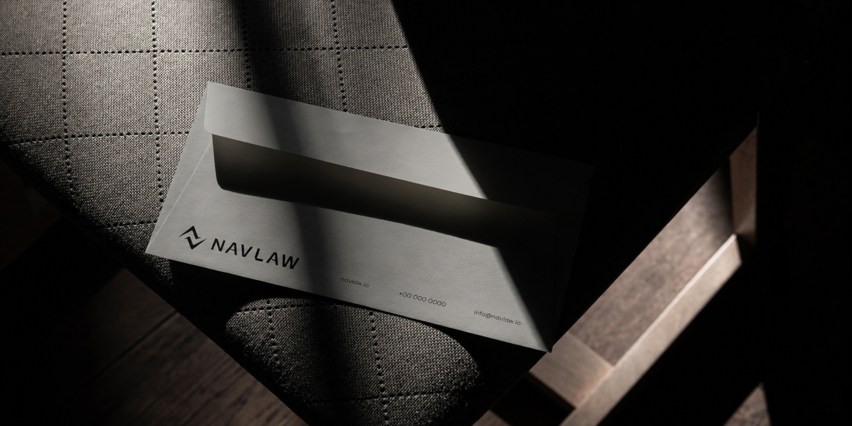 NAVLAW premium correspondence envelope — metallic brushed stock with NAVLAW combination mark, contact details showing navlaw.io and info@navlaw.io, photographed on dark woven fabric with dramatic lighting